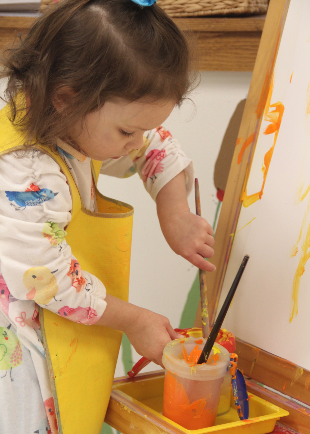 Farmington Valley Academy Montessori - Toddler Assistant. The toddler is preparing to create a work of art. She is focusing on filling her brush with yellow and orange paint as she stands at an easel.