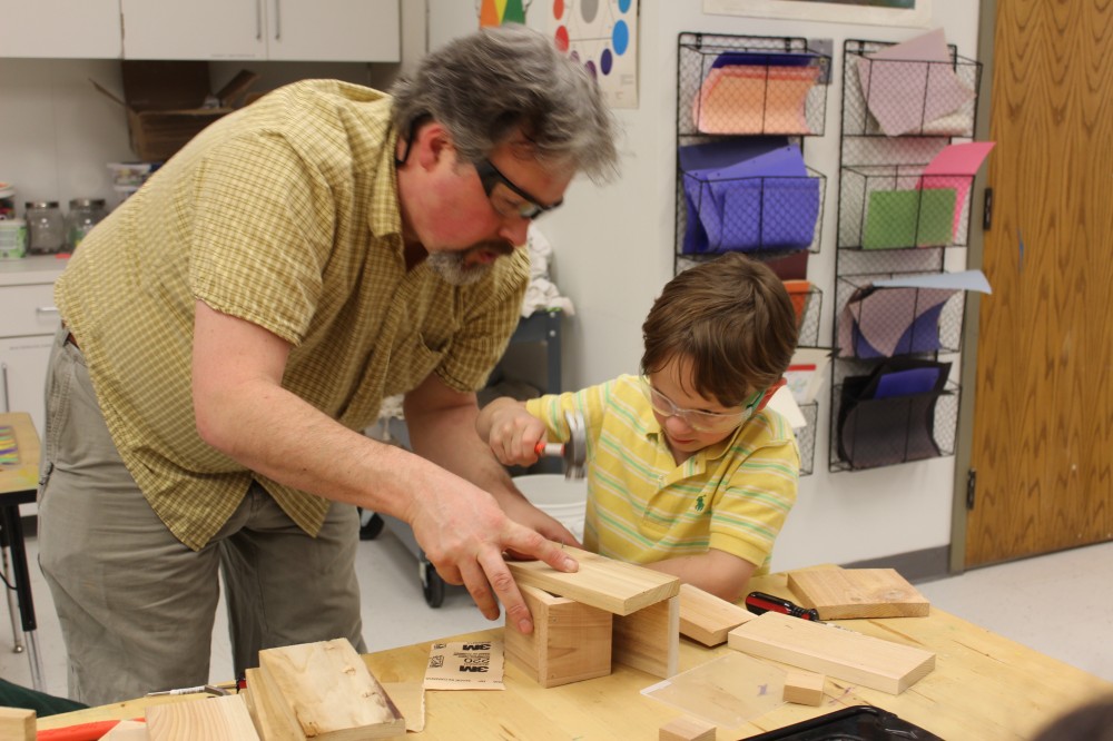 Farmington Valley Academy Montessori - Primary Associate Teacher. The young boy is working, completing a woodwork project. The assistant is holding 2 pieces of wood in place, as the boy, strikes a nail with a hammer, to join the 2 pieces together. The practical life experience is very important at this age.
