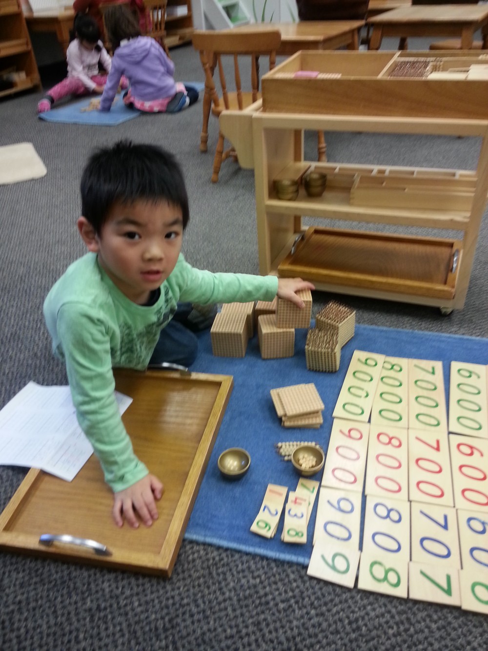 Farmington Valley Academy Montessori - Primary Associate Teacher. The young boy is working, on the mat, with the Golden Bead material, and the large number cards. It looks like he is being introduced to the decimal system.