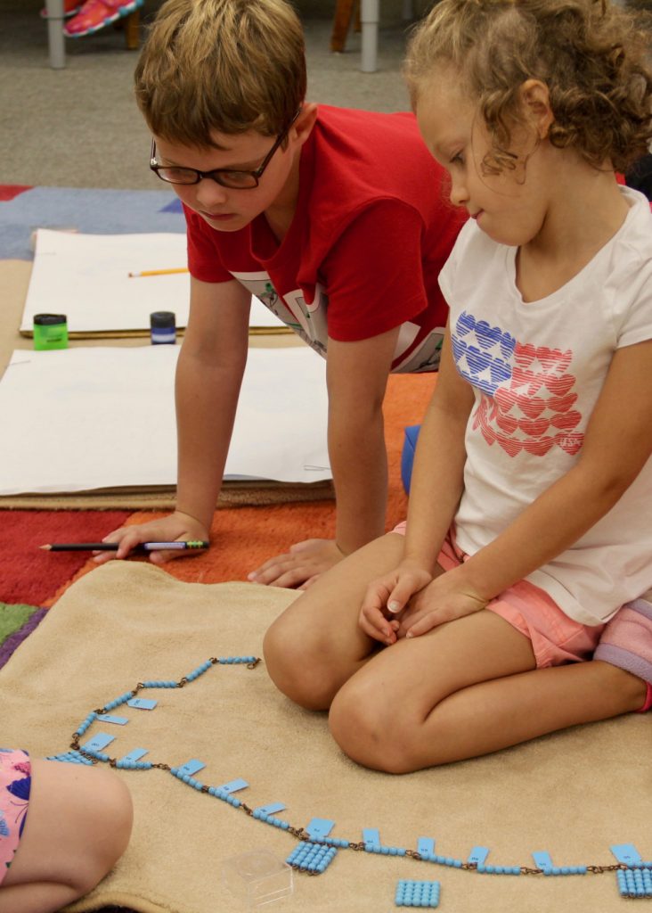 Farmington Valley Academy Montessori - Primary Associate Teacher. Collaborative work is an important aspect to learning with different age groups. These 2 young pre-schoolers are trying to work out the skip and count mathematics activity. Blue beads on the floor mat with a young girl in the bead circle, and the boy leaning over to try and explain something to her.