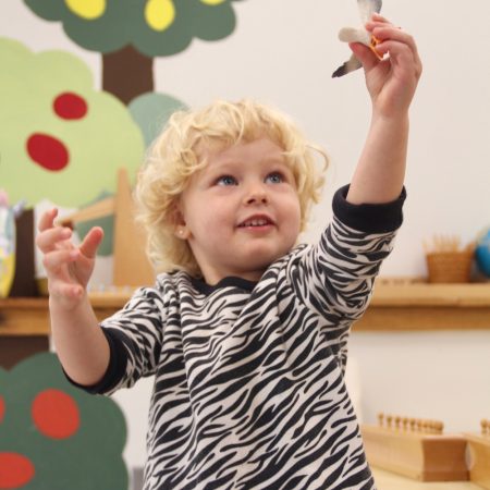 Farmington Valley Academy Montessori - Toddler Assistant. The toddler is "flying" a plastic bird, imagining what it would feel like to be a bird, free to fly anyway.